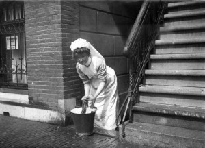 Dienstbode [dienstmeid, huishoudster] in Amsterdam bezig met het dweilen van de stoep. Amsterdam, 1912.